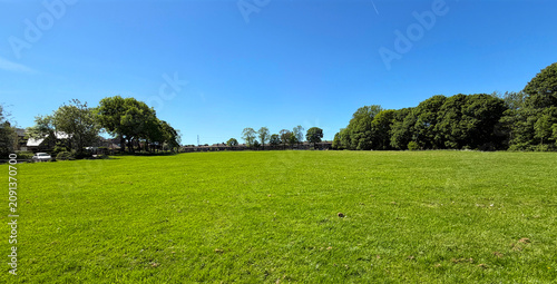 Photography A wide, open grassy field spreads out beneath a clear blue sky, with trees lining the edges and casting soft shadows across the vibrant green grass in Shelf Hall Park, Shelf, Bradford, UK