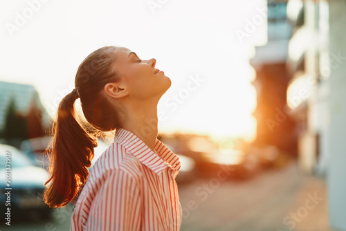 Bild auf Leinwand Woman portrait tilts her head back, gazing at the sunset over an urban street, sunlight kissing her striped shirt and ponytail in warm glow