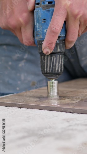 Hands operating blue cordless drill with hole saw attachment pressed into wood board on work surface, showing firm grip, dusty skin, worn tool body and precise carpentry work in