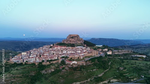 Aerial view of Morella's hilltop castle and walled medieval town at dusk, full moon rising over the surrounding mountains of Els Ports, Castellon, Spain