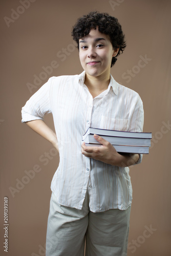 Young adult woman holding a stack of books on one arm. Vertical studio portrait with copy space on brown background. Ideal for university student, adult education, teaching, reading, and writing.