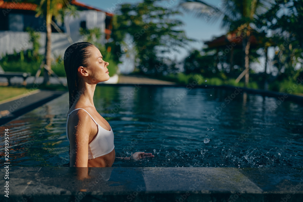 Fototapeta premium Young woman with wet hair enjoys a peaceful moment in a swimming pool surrounded by tropical greenery during a sunny day.