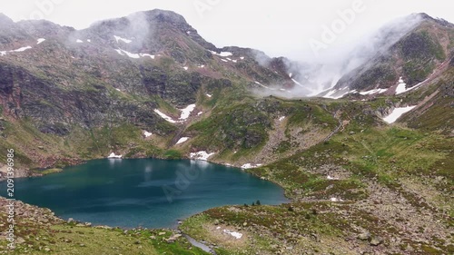 Aerial view of a deep turquoise glacial lake nestled among rocky alpine slopes with snowmelt streams, patches of snow, and low clouds rolling over the Tristaina Lakes, Andorra, Pyrenees