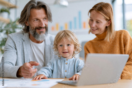 Happy parents smile while sitting with their child at a laptop. Child looks curiously at the screen with bright eyes. Parent gestures toward the laptop, pointing to something on the screen