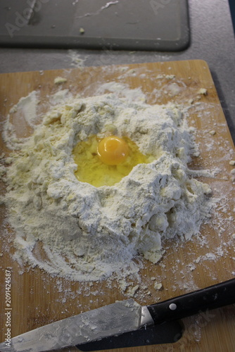 Mound of flour with a cracked raw egg in the center on a wooden board, knife positioned nearby, kitchen countertop seen in the background