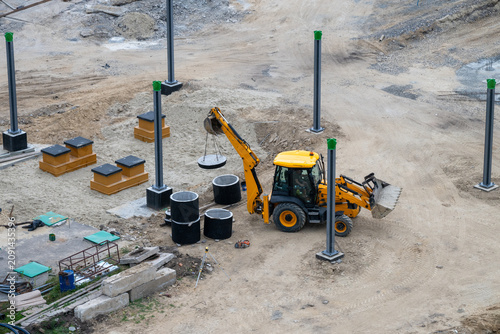 Cuadro en lienzo a yellow backhoe loader using its rear arm to carefully lift and position a concrete manhole cover among scattered building materials and installed metal pillars