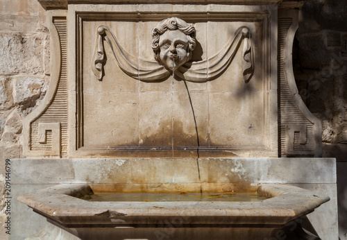 A decorative stone wall fountain with a carved cherub face spouting water into a marble basin in Valletta, Malta