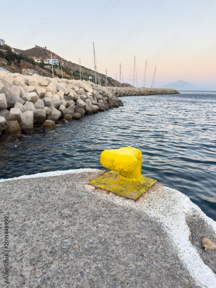 Fototapeta premium Yellow mooring bollard beside calm Aegean harbor at dusk in Agios Kirikos, Ikaria island, Greece