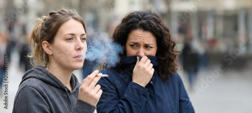 One woman stands smoking a cigarette on a busy street while another woman shows discomfort from the smoke. People walk by in the background during the day