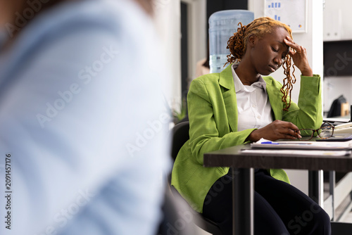 Fotografie Black female employee feeling unmotivated, struggling with ongoing business operations while tired businesswoman rests hand on forehead, showing exhaustion from continuous workload and unending tasks