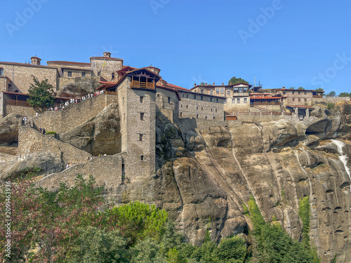 Kalambaka, Greece - July 24, 2024: Meteora, Holy Monastery of the Great Meteoron. Look upon the monastery buildings from the nearby rock plateau, separated by a deep ravine under a blue sky. Green fol