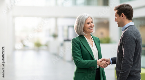 Senior Businesswoman Shaking Hands with Male Colleague for Successful Partnership Deal or Mentorship Greeting in Bright Modern Corporate Office Lobby Background with Copy Space