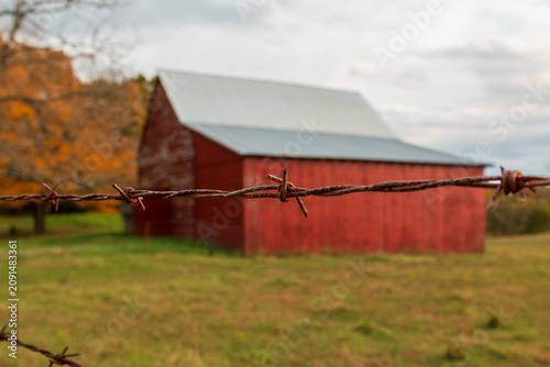 Obraz na plátně A shallow depth of field image of a prong on a barbed wire fence with a blurry red barn and trees with autumn leaves in the background
