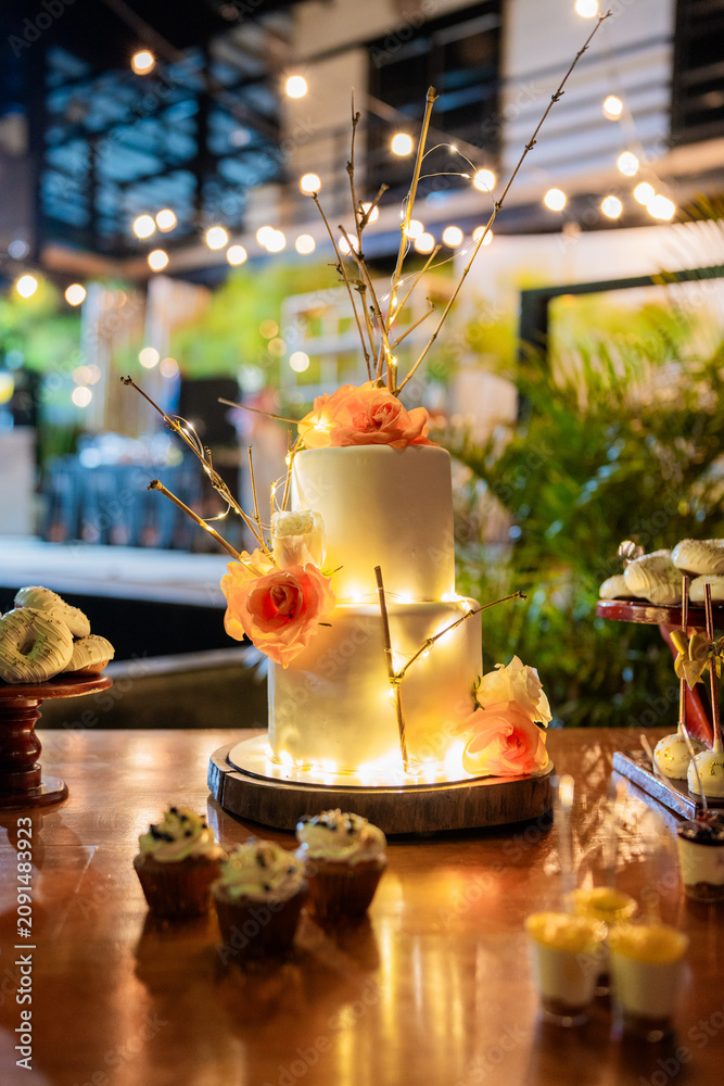 Fototapeta premium Elegante pastel de dos pisos decorado con rosas y luces para celebración de boda, cumpleaños o evento especial