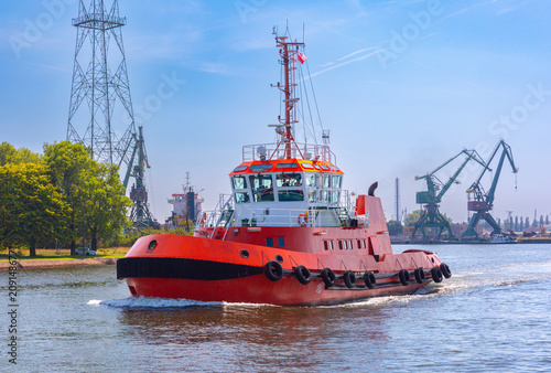Red harbor tugboat navigating a waterway near the Gdansk Shipyard with industrial cranes and port infrastructure in Gdansk, Poland.