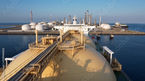 Aerial view of an LNG carrier bow with a spherical gas tank approaching a coastal natural gas terminal, gas tanker mooring at a liquefied natural gas plant by the sea