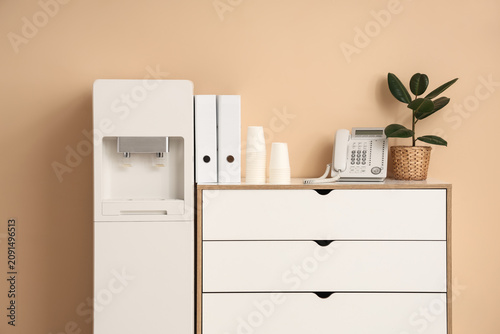 Modern water cooler and chest of drawers near beige wall