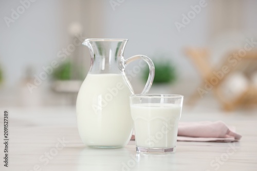 Fresh milk in glass and jug on light table indoors