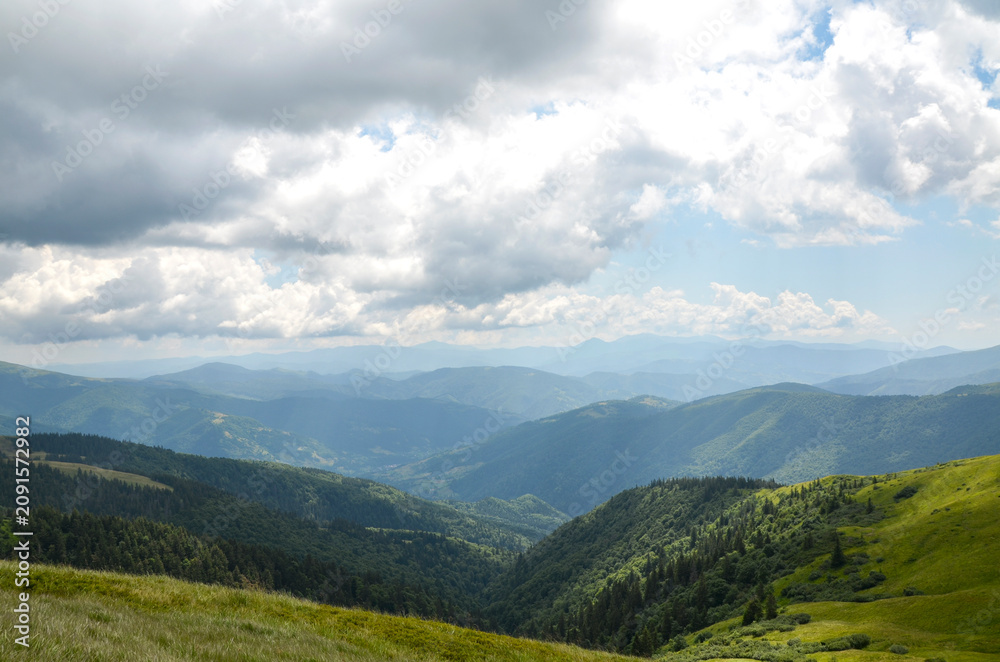 Fototapeta premium Dramatic sunlight and cloud shadows sweep across lush green ridges and vast, rolling mountain ranges stretching toward the horizon under a heavy, overcast sky. Carpathian Mountains, Ukraine 