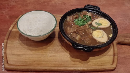 A comforting meal of braised pork belly with soy-marinated eggs served in a clay pot alongside steamed white rice, presented on a wooden serving board.
