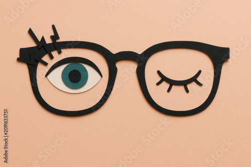 Playful paper cut glasses craft with one open eye and one closed eye winking, teal iris, isolated on peach background, fun cartoon optical art