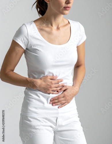 A woman in white clothing, clutching her abdomen with both hands, expressing distress and discomfort. She appears to be suffering from abdominal pain