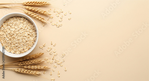 Raw oatmeal in white bowl with wheat ears, healthy breakfast ingredients on beige background, organic oat flakes flat lay with copy space