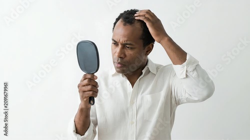 A Black man seriously worries about thinning hair on the top of his head and at his hairline, using a hand mirror.