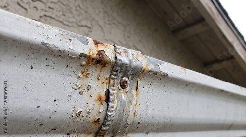 Rusty metal rain gutter close up, with peeling white paint and corroded seam fastener, hanging on house exterior eaves, property maintenance