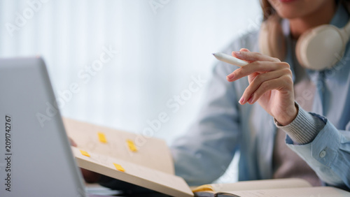 Young Asian woman holding pen while studying with laptop and notebook at desk with headphones with copy space