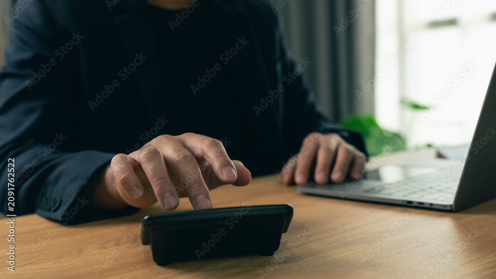Fototapeta premium Businessman using calculator for financial calculation with laptop on wooden desk in modern office interior background