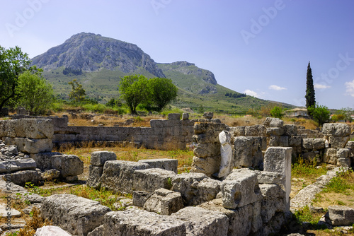 Ancient ruins of Corinth, Greece, with a statue fragment in the foreground and a mountain in the background under a clear blue sky.