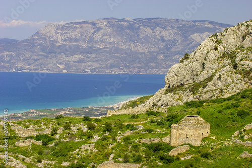 Ancient ruins of a stone structure on a grassy hillside overlooking the blue sea and distant mountains under a clear sky.