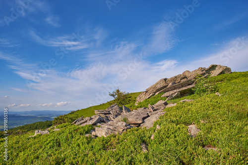 A bright blue sky with wispy clouds stretches over a vibrant green mountain slope scattered with rugged rock formations in beautiful wilderness landscape