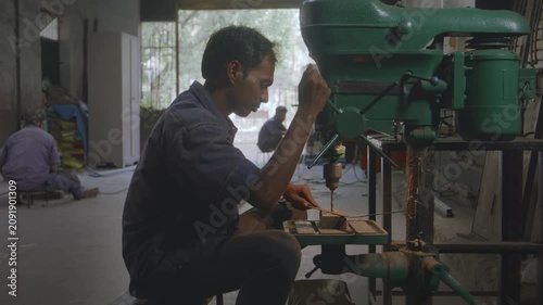 A close up indoor shot of an Indian Asian male worker drilling a hole in a metal rod with precision using bench mounted drill press machine, coworkers working in the background in fabrication workshop