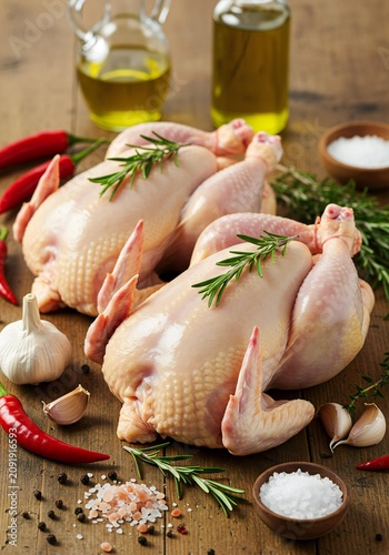 Two whole raw chickens seasoned with rosemary, garlic, chili, and olive oil on a wooden table