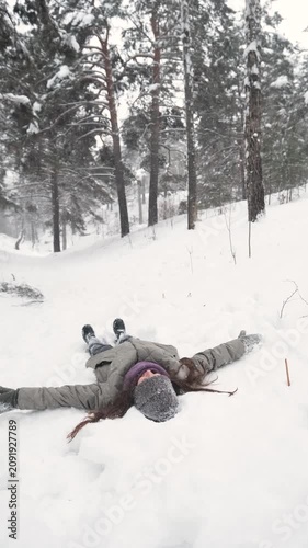 A beautiful young woman is making a snow angel in a snow-covered forest. She is happily spending her free time and enjoying the first snow. An active winter pastime.