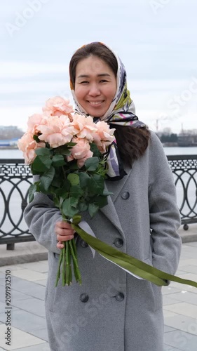 A beautiful woman wearing a headscarf stands on a city street in early spring. She smiles and holds a large bouquet of pink roses.