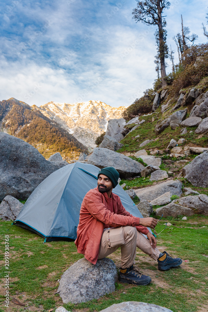 Fototapeta premium Man sitting on a rock near a camping tent at Kareri Lake in the mountains of Himachal Pradesh