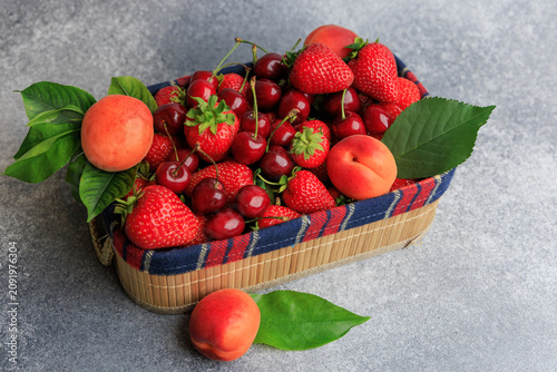 Fresh strawberries cherries and apricots in a woven basket on a gray surface during sunny weather