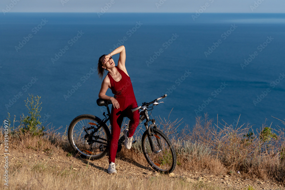 Fototapeta premium Cyclist woman nature, athletic female wearing red sportswear posing with bicycle on a cliff above the blue sea at sunny day