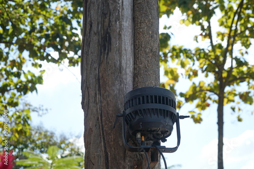 A black outdoor landscape spotlight or PAR Light (Parabolic Aluminized Reflector)  is mounted on a textured tree trunk with green and yellow leaves around, illuminated by daylight under a partly cloud