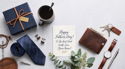 Father's day gift arrangement featuring a blue gift box, leather wallet, necktie, wristwatch, keys, coffee mug, and a greeting card on a clean white textured background for holiday celebration