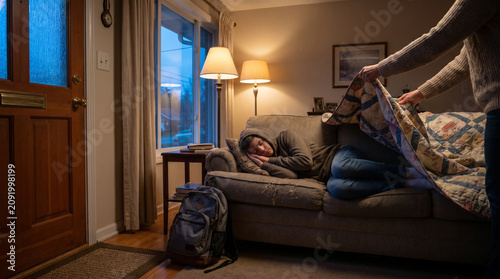 Parent Gently Covering Sleeping Teenager with Blanket on Couch After School, Warm Lamp Light and Blue Dusk Through Window