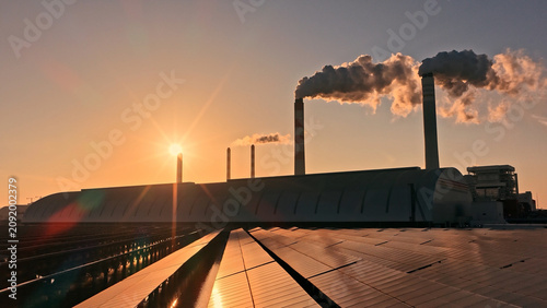 Solar sun panel field near fossil fuel power plant. Transition traditional coal energy to renewable electricity. Industrial landscape contrast between clean tech and pollution. Factory smoking chimney