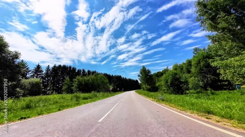 Winding asphalt road bordered by lush green trees and vibrant blue sky full of clouds