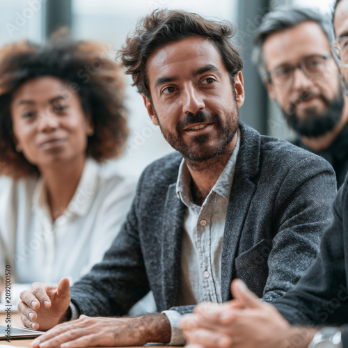 Diverse professionals engaged in a focused business meeting. Confident man gestures while speaking, eyes directed forward. Colleagues listen attentively, showing engagement and professionalism