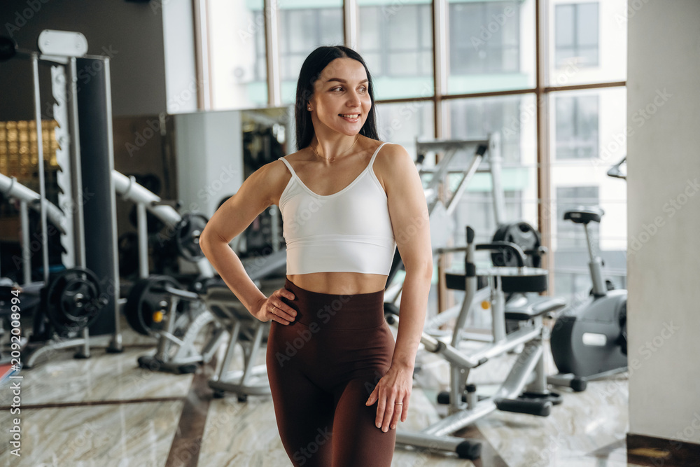 Obraz premium Female fitness enthusiast in white sports bra and brown leggings poses confidently in a modern gym with exercise equipment and large windows in the background