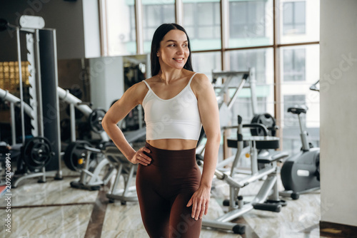 Female fitness enthusiast in white sports bra and brown leggings poses confidently in a modern gym with exercise equipment and large windows in the background