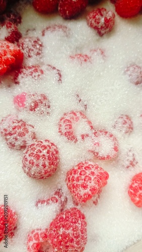 Extreme close-up of vibrant red raspberries scattered on a bed of white granulated sugar, highlighting their texture and seeds.